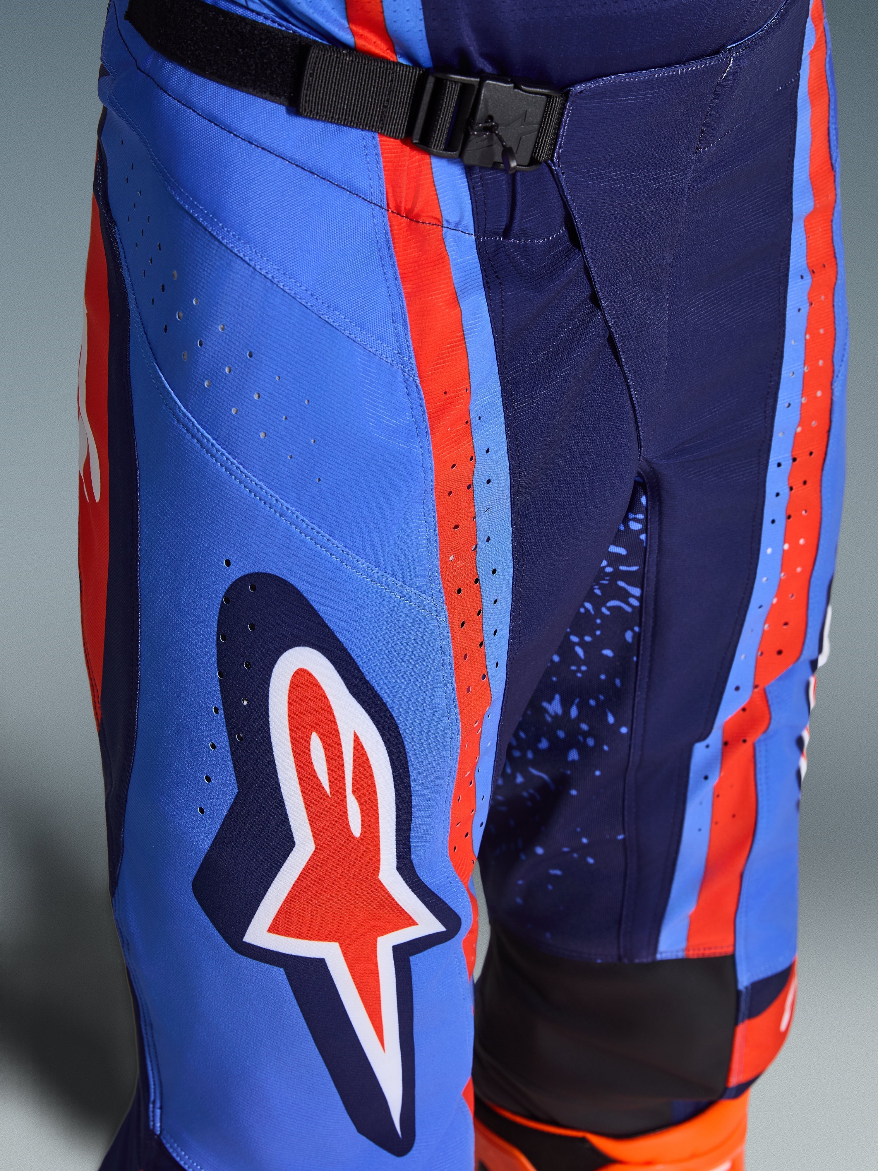Alpinestars Techstar Nomur Pants 2026, pantaloni da motocross in blu navy scuro, arancione e blu UCLA, primo piano che mostra la chiusura in vita con fibbia magnetica SX2, perforazioni laser sulla coscia e l'iconico logo a stella, equipaggiamento da fuoristrada resistente.