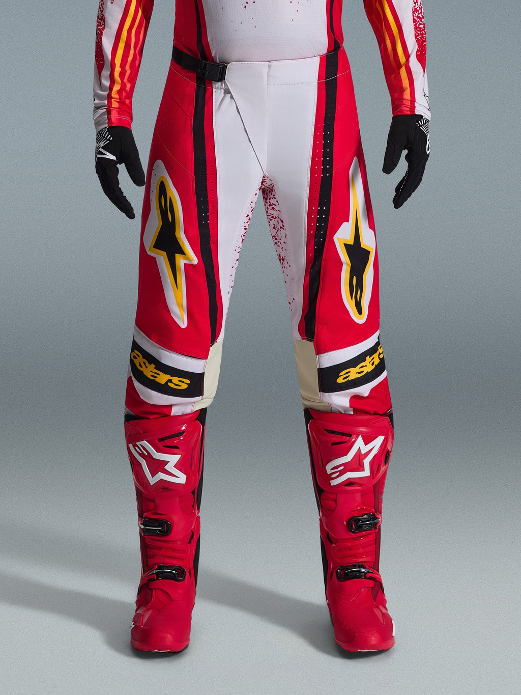 Alpinestars pantaloni Techstar Nomur 2026, pantaloni da motocross, rosso e bianco con accenti neri e gialli, indossati con stivali rossi coordinati e guanti neri, abbigliamento tecnico per gare off-road