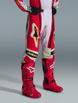 Alpinestars 2026 Techstar Nomur Pants, pantaloni da motocross, rosso, bianco e nero con accenti gialli, indossati con stivali da motocross rossi coordinati e guanti neri, con logo Alpinestars giallo e marchio "astars" sulle ginocchia, abbigliamento professionale per corse fuoristrada