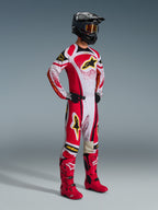 Pantaloni Alpinestars 2026 Techstar Nomur, pantaloni da motocross, rosso e bianco con accenti neri e gialli, indossati da un pilota con maglia coordinata, stivali rosso acceso e casco nero con occhiali dorati, abbigliamento professionale completo da competizione off-road