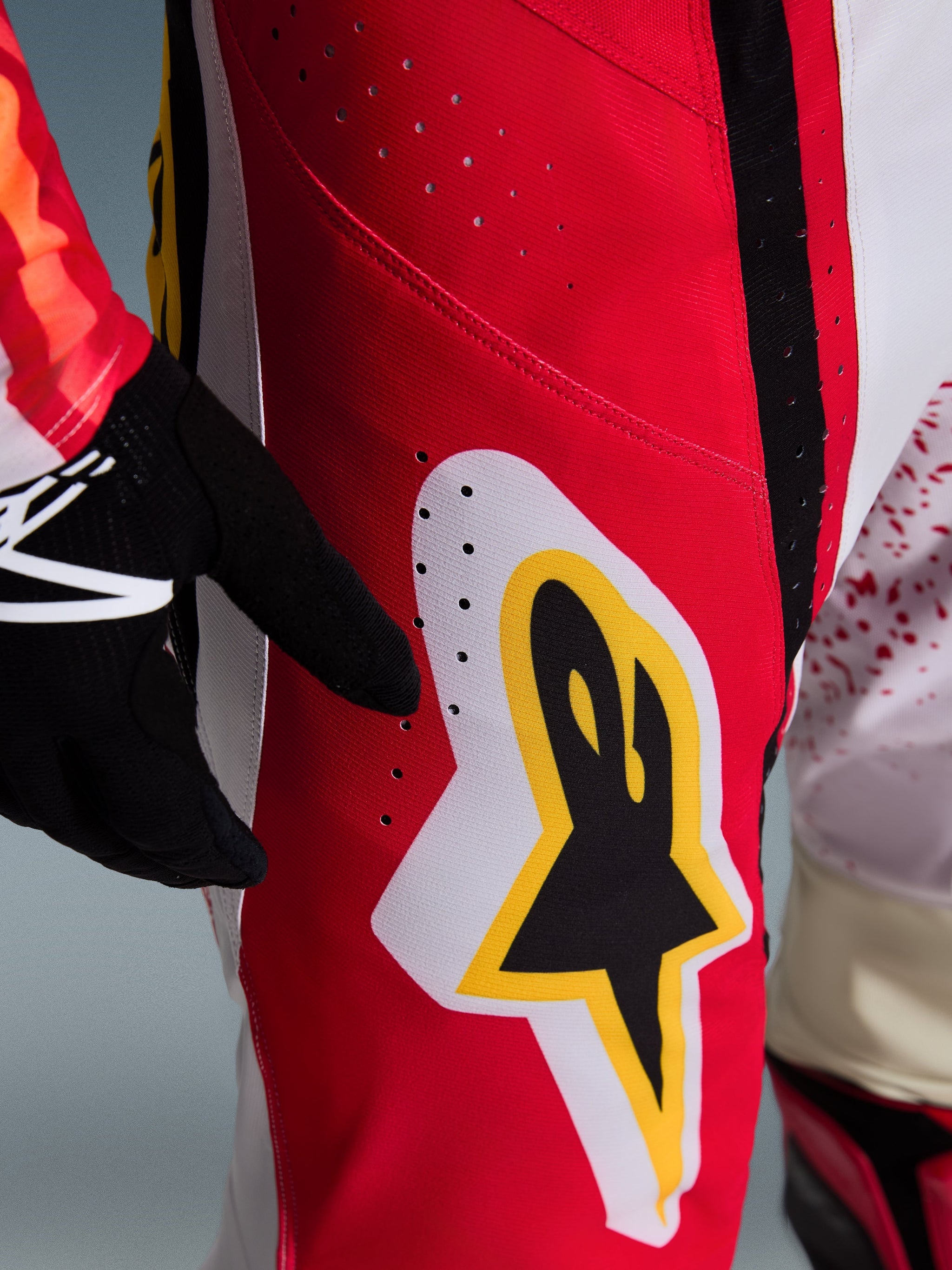 Alpinestars 2026 Techstar Nomur Pants, pantaloni da motocross, rosso con accenti bianchi e gialli, primo piano della zona coscia con fori di ventilazione tagliati al laser e grande logo a stella, abbinati a guanti neri e maglia colorata per prestazioni off-road