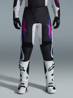 Pantaloni Alpinestars 2026 Fluid Grid, pantaloni da motocross neri con design a stelle viola e blu, indossati con stivali da motocross bianchi e guanti neri, caratterizzati da pannelli rinforzati e vestibilità sportiva off-road, sfondo grigio