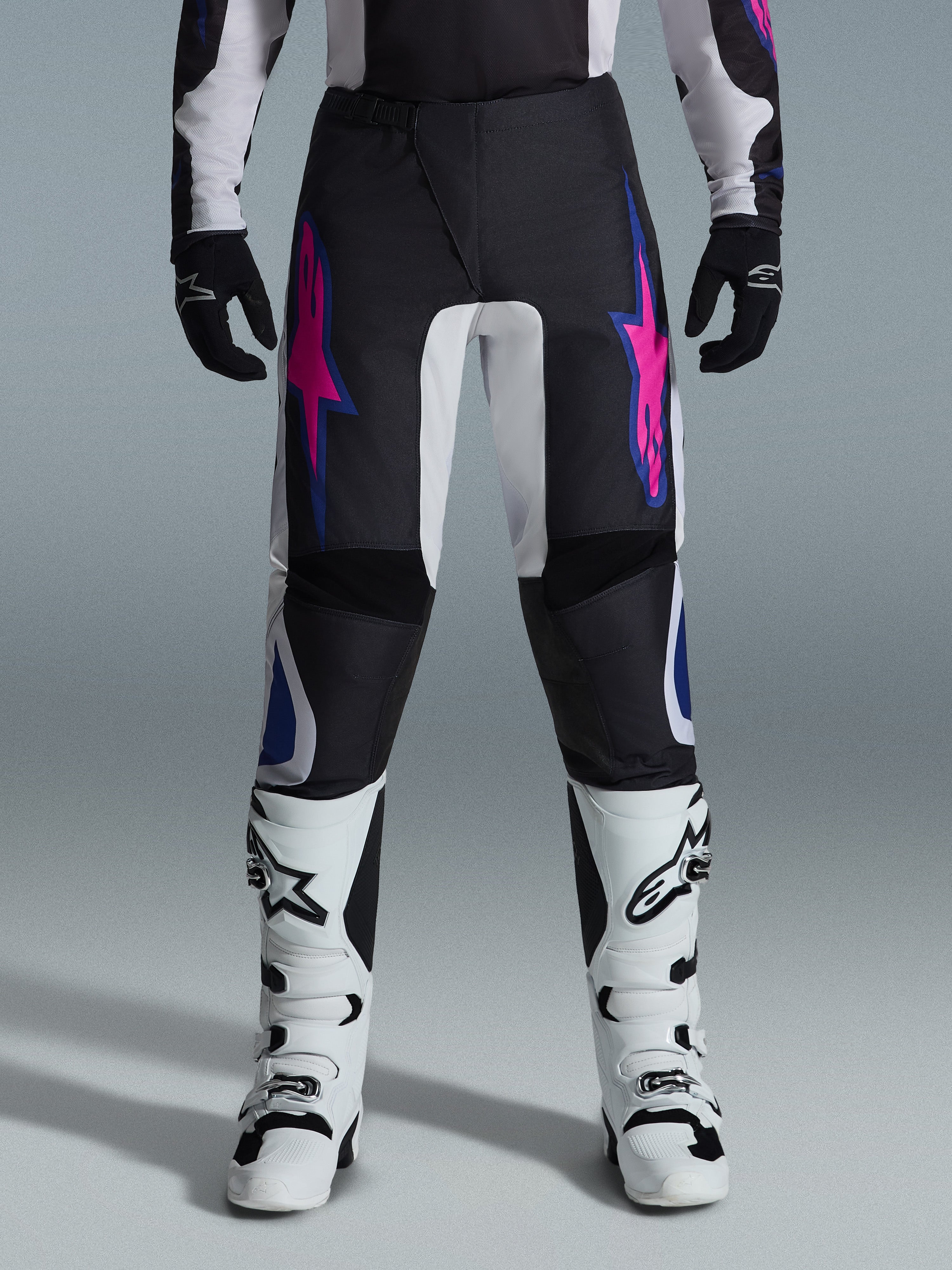 Pantaloni Alpinestars 2026 Fluid Grid, pantaloni da motocross neri con design a stelle viola e blu, indossati con stivali da motocross bianchi e guanti neri, caratterizzati da pannelli rinforzati e vestibilità sportiva off-road, sfondo grigio