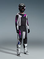 Pilota di motocross con abbigliamento completo da gara, pantaloni Alpinestars 2026 Fluid Grid in Nero Grigio Chiaro e Viola, maglia coordinata con grafiche rosa e blu, casco e occhiali neri, stivali bianchi e neri e guanti neri, completo coordinato per corse fuoristrada