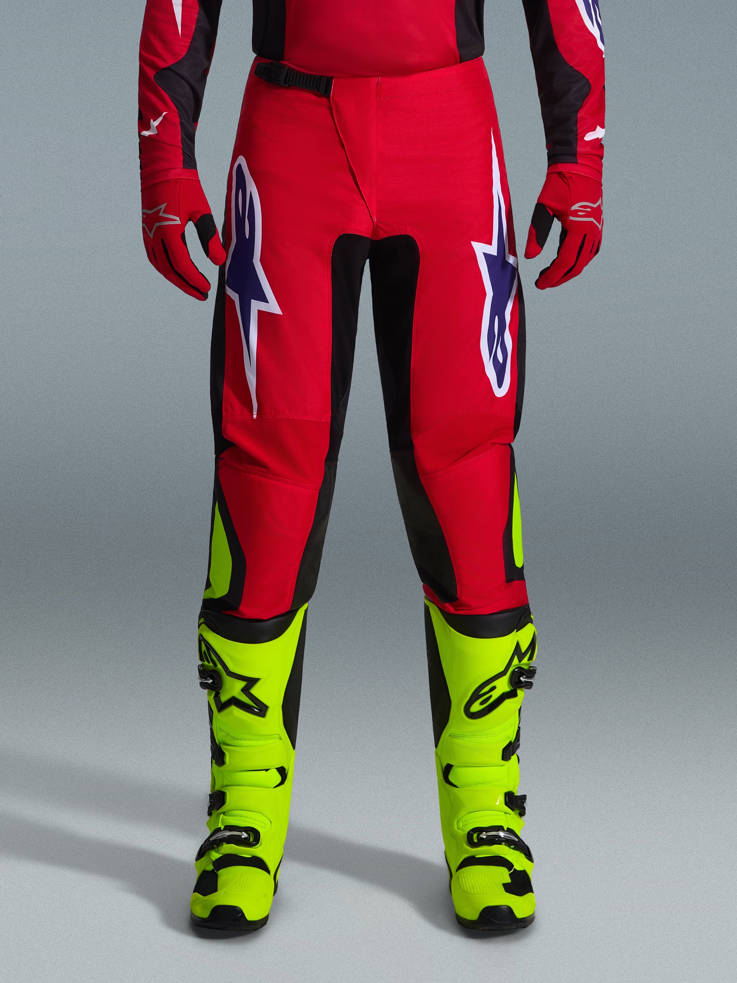 Alpinestars pantaloni Fluid Grid 2026, pantaloni da motocross, rosso con pannelli neri e accenti giallo neon, grandi loghi a stella bianchi e viola sulle cosce, indossati con guanti rossi e stivali giallo neon, costruzione resistente per gare off-road