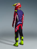 Alpinestars 2026 Fluid Grid Pants, pantaloni da motocross, rosso, nero e viola, indossati da un pilota visto da dietro con maglia coordinata e stivali giallo fluo, con branding audace e design ergonomico per prestazioni off-road.