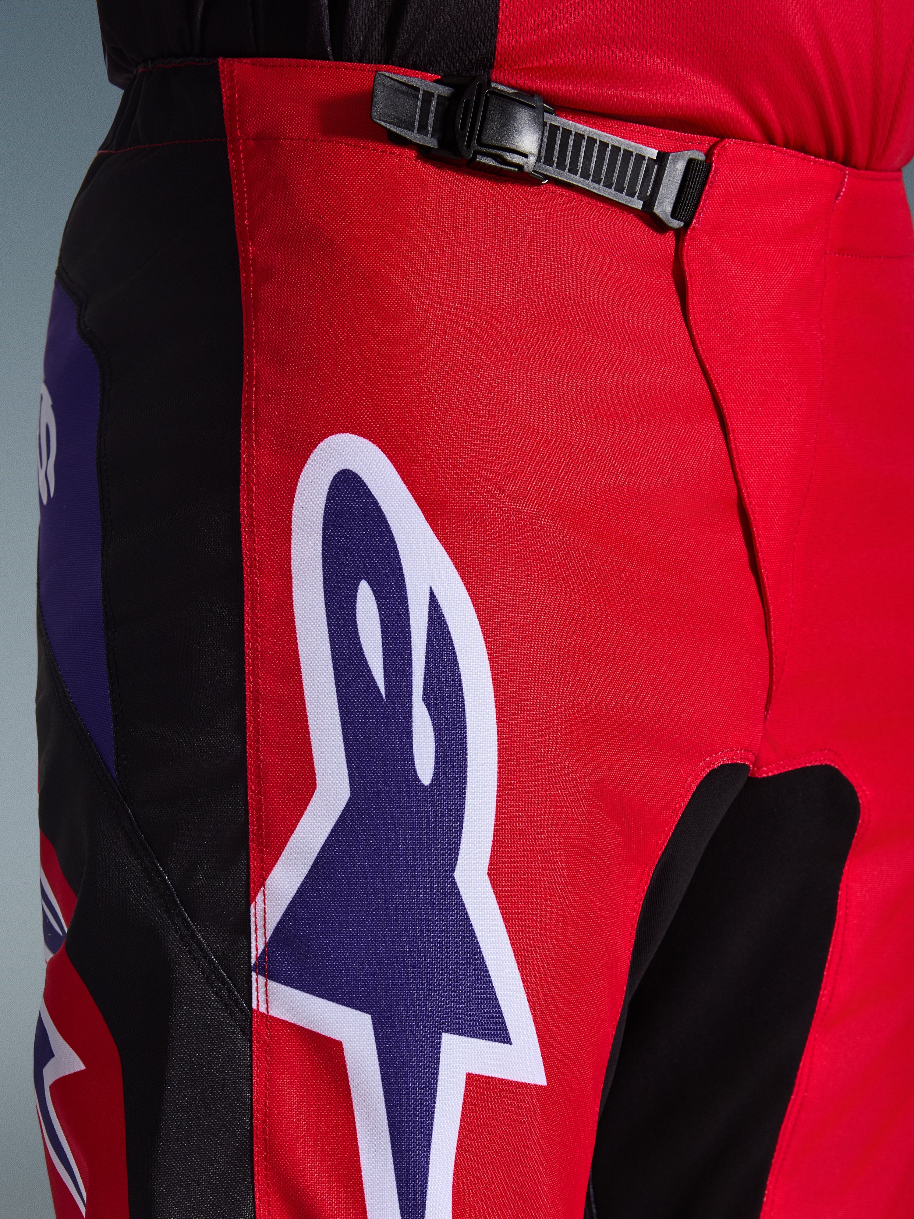 Pantaloni Alpinestars Fluid Grid 2026, pantaloni da motocross, rosso nero e viola, primo piano della zona della vita e dei fianchi con sistema di chiusura a cricchetto nero e grande logo Alpinestars a stella viola con bordo bianco, tessuto strutturato resistente per corse off-road