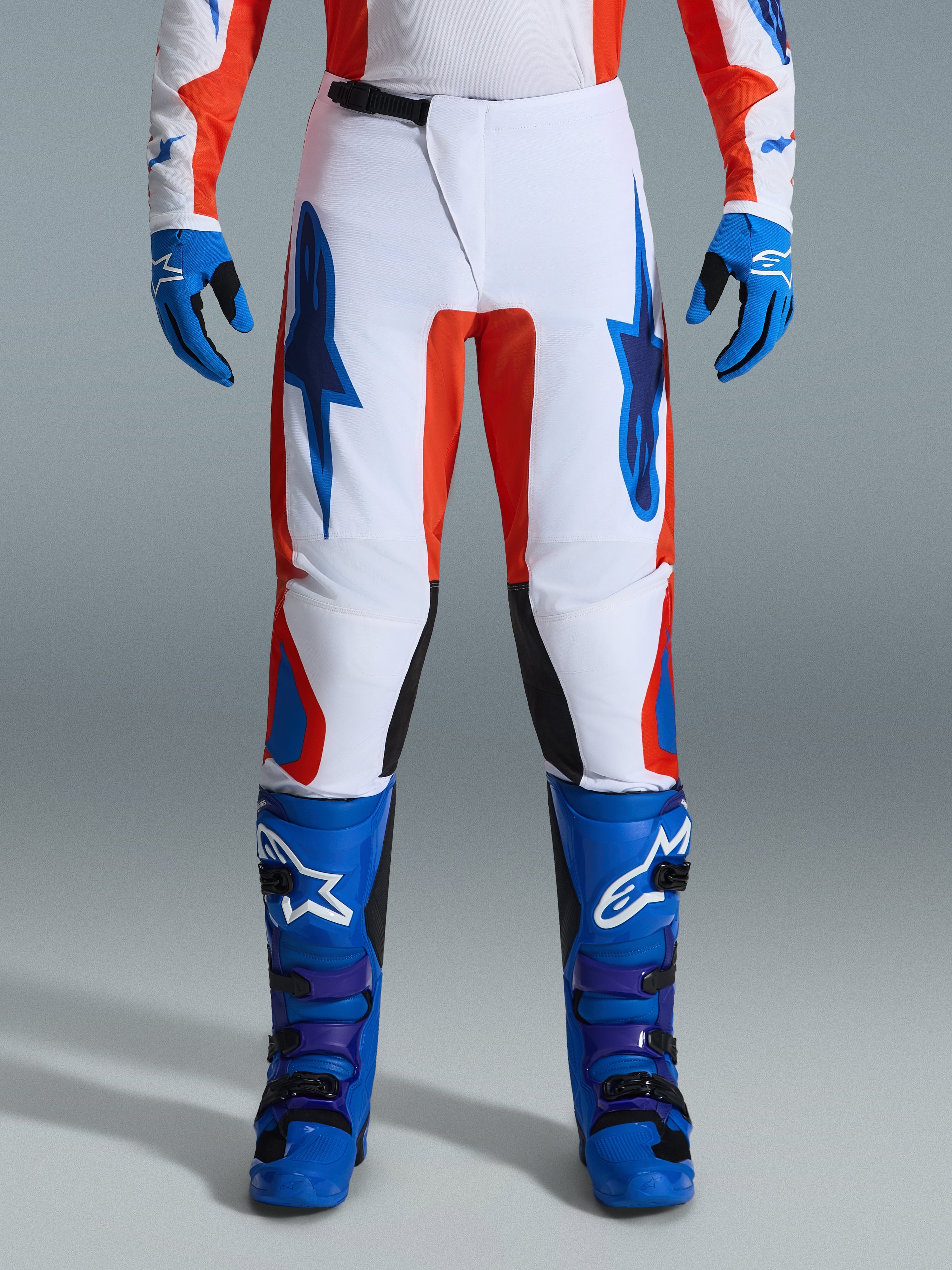 Alpinestars 2026 Fluid Grid Pants, pantaloni da motocross, arancione e blu UCLA, indossati con stivali e guanti blu coordinati, caratterizzati da telaio bianco con loghi blu audaci e accenti arancioni, design ergonomico per prestazioni fuoristrada