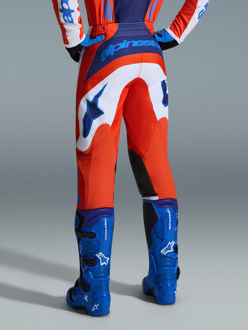 Alpinestars 2026 Fluid Grid Pants, pantaloni da motocross, arancione e blu UCLA con pannelli bianchi, vista posteriore con logo blu sulla seduta e logo a stella, abbinati a stivali da motocross blu coordinati, equipaggiamento professionale per fuoristrada