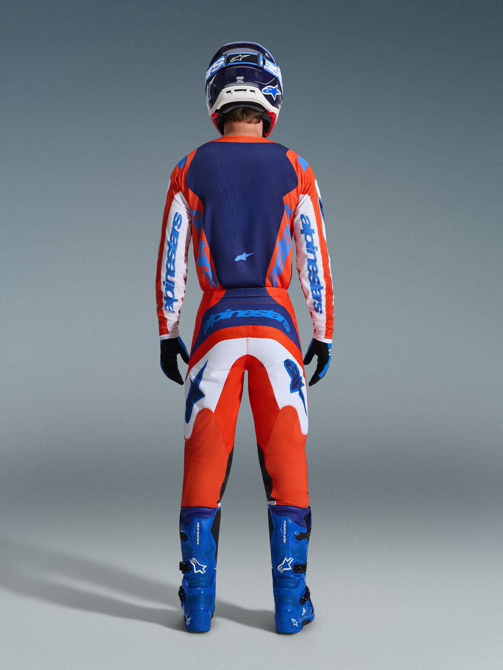 Alpinestars 2026 Fluid Grid Pants, pantaloni da motocross, arancione e blu UCLA, vista posteriore completa di un pilota che indossa un set da gara completo con maglia coordinata, stivali blu e casco, con branding prominente e pannelli ergonomici per la guida fuoristrada.
