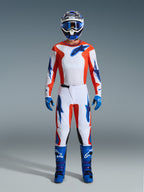 Pilota di motocross con abbigliamento completo da gara, pantaloni Alpinestars 2026 Fluid Grid in arancione e blu UCLA, maglia bianca con maniche arancioni e loghi blu, casco e occhiali blu, stivali e guanti blu, completo coordinato per corse fuoristrada