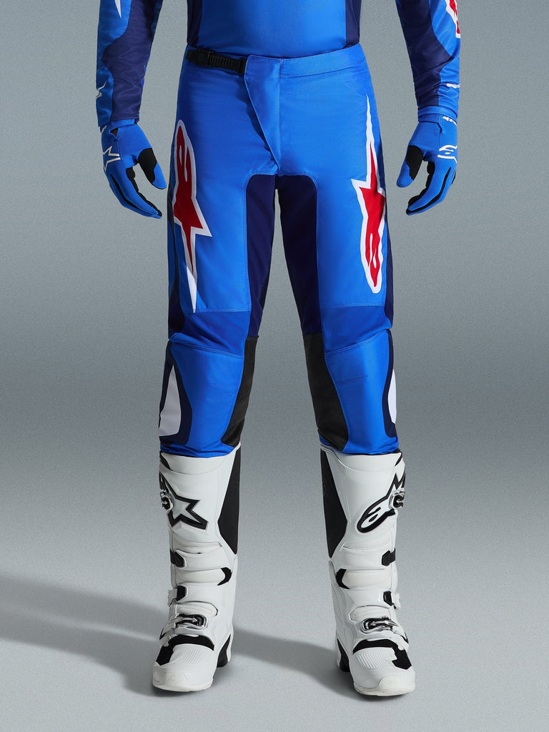 Alpinestars 2026 Fluid Grid Pants, pantaloni da motocross, blu e rosso, con vistosi loghi a stella rossi e bianchi sulle cosce, indossati con guanti blu coordinati e stivali bianchi da fuoristrada, vestibilità atletica con pannelli resistenti per le gare.