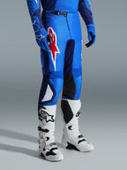 Alpinestars 2026 Fluid Grid Pants, pantaloni da motocross, blu e rosso, indossati con stivali da motocross bianchi e guanti blu, con un grande logo a stella rosso sulla coscia e pannelli interni neri sulle ginocchia, progettati per la guida fuoristrada