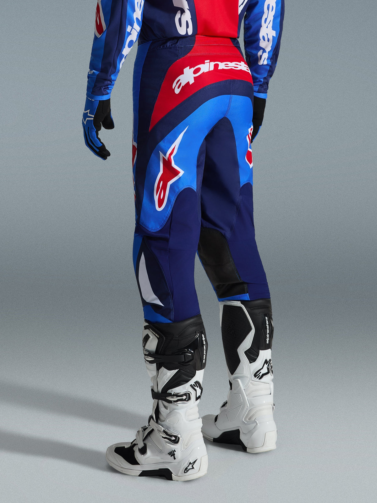 Pantaloni Alpinestars Fluid Grid 2026, pantaloni da motocross, blu e rosso, vista posteriore con branding Alpinestars e design ergonomico, indossati con stivali Tech 7 bianchi e neri e maglia coordinata, attrezzatura ad alte prestazioni per il fuoristrada