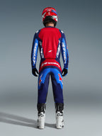 Pilota di motocross visto di spalle con abbigliamento completo da gara, Pantaloni Fluid Grid 2026 in Blue Red, maglia coordinata rossa e blu con logo Alpinestars, casco rosso e blu, stivali bianchi e neri, completo coordinato per il fuoristrada