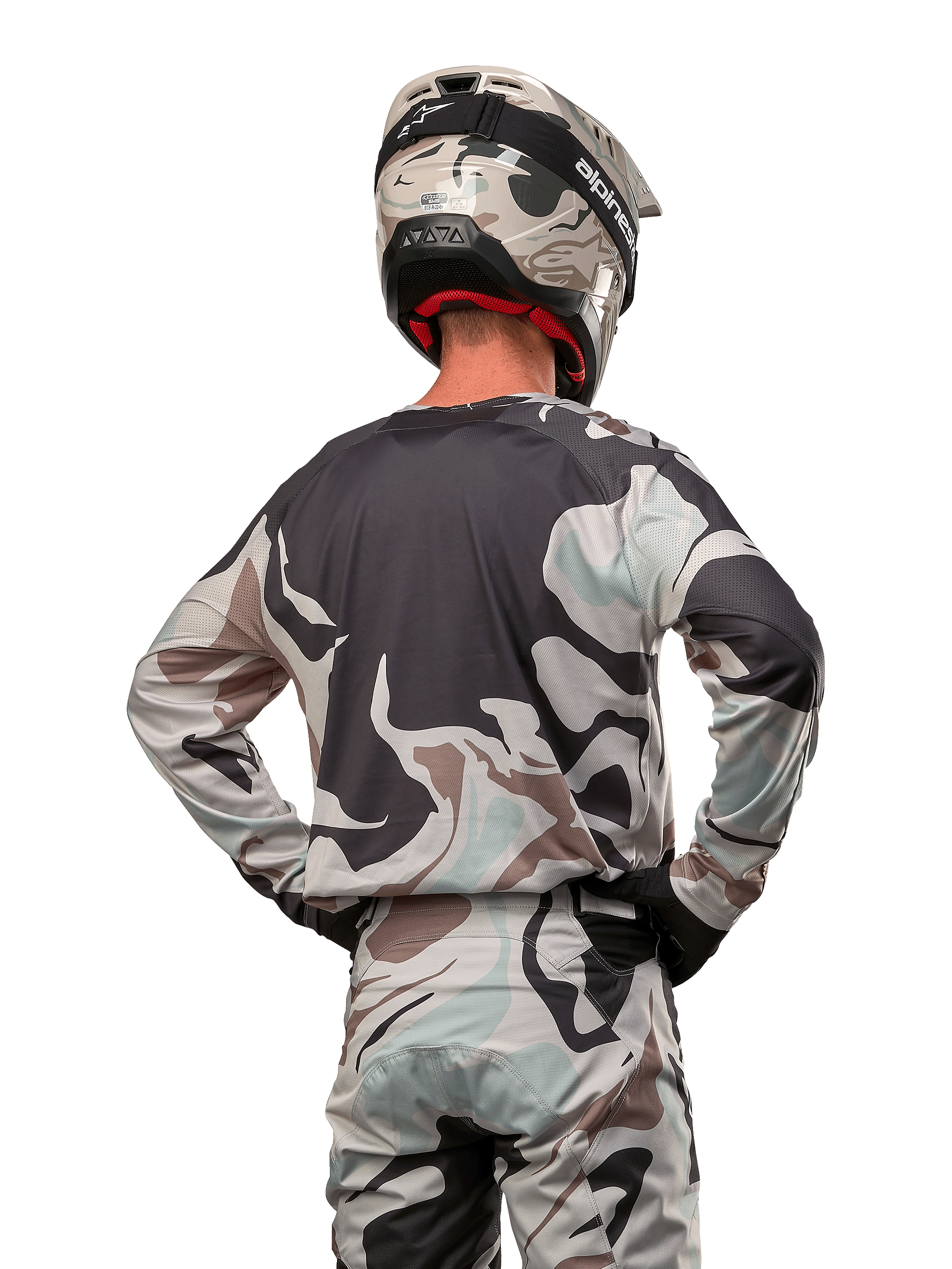 Pilota di motocross visto da dietro che indossa la maglia Alpinestars Racer Tactical 2024 in Iron Camo Dust Gray, pantaloni e casco mimetici coordinati, mani sui fianchi, abbigliamento da gara fuoristrada coordinato.