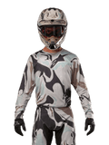 Alpinestars Maglia Racer Tactical 2024, maglia da motocross, motivo mimetico Iron Camo Dust Gray, pilota che indossa casco e occhiali coordinati con guanti neri, abbigliamento tecnico per fuoristrada