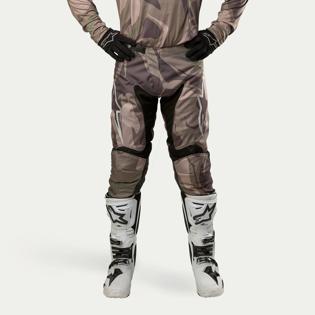 Pantaloni Alpinestars Racer Tactical 2024, pantaloni da motocross, mimetico verde militare e marrone, con grandi loghi Alpinestars bianchi sulle cosce e pannelli interni neri, indossati con stivali da motocross bianchi e neri, guanti neri e maglia mimetica coordinata, vista frontale