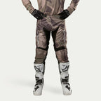 Pantaloni Alpinestars Racer Tactical 2024, pantaloni da motocross, mimetico verde militare e marrone, con grandi loghi Alpinestars bianchi sulle cosce e pannelli interni neri, indossati con stivali da motocross bianchi e neri, guanti neri e maglia mimetica coordinata, vista frontale