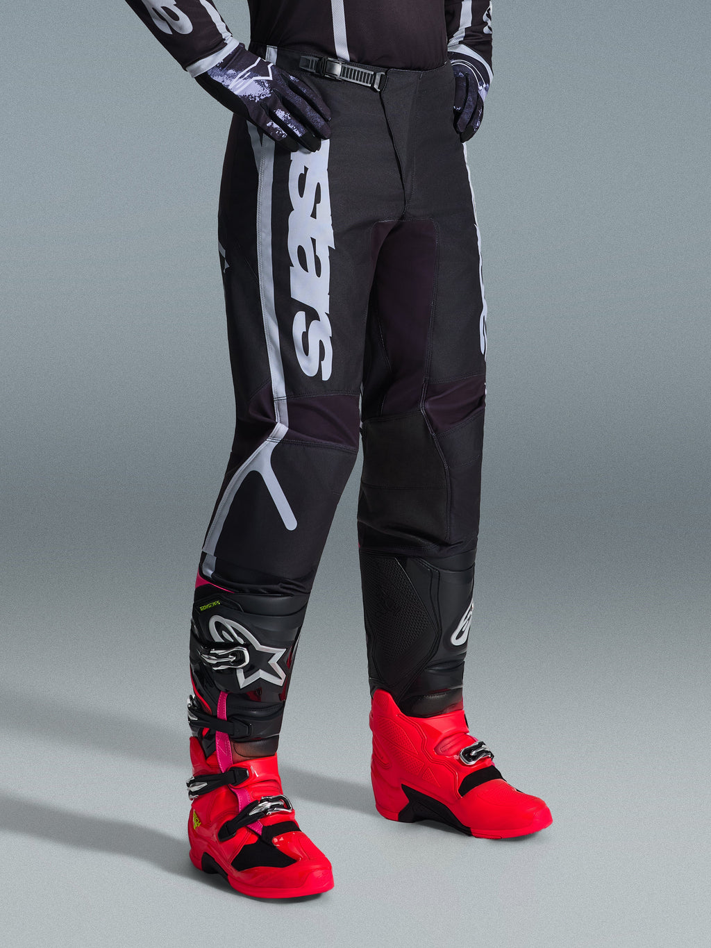 Pantaloni Alpinestars 2026 Fluid Apex, pantaloni da motocross, nero e grigio, indossati con stivali rosa vivace e rossi e maglia abbinata nera e bianca, con logo Alpinestars verticale sulla coscia, pannelli rinforzati e chiusura a cricchetto in vita per prestazioni fuoristrada.