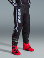 Pantaloni Alpinestars 2026 Fluid Apex, pantaloni da motocross, nero e grigio, indossati con stivali rosa vivace e rossi e maglia abbinata nera e bianca, con logo Alpinestars verticale sulla coscia, pannelli rinforzati e chiusura a cricchetto in vita per prestazioni fuoristrada.