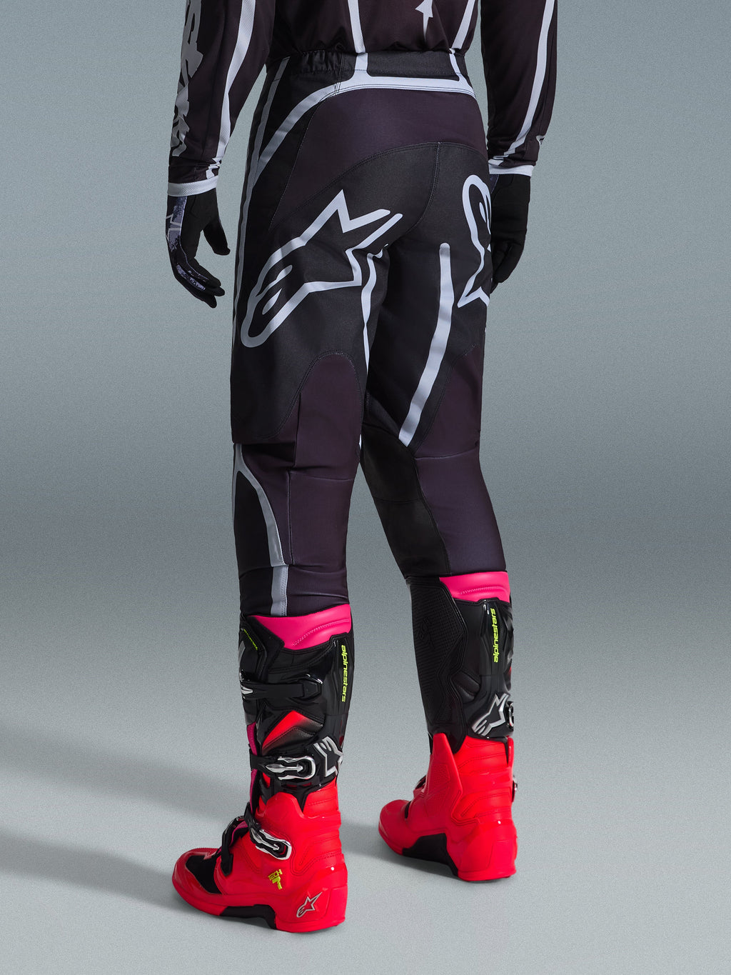 Alpinestars Pantaloni Fluid Apex 2026, pantaloni da motocross, nero e grigio, vista posteriore con grandi loghi Alpinestars bianchi sulla seduta, abbinati a stivali rosso acceso e rosa, costruzione in poliestere 600D per prestazioni off-road
