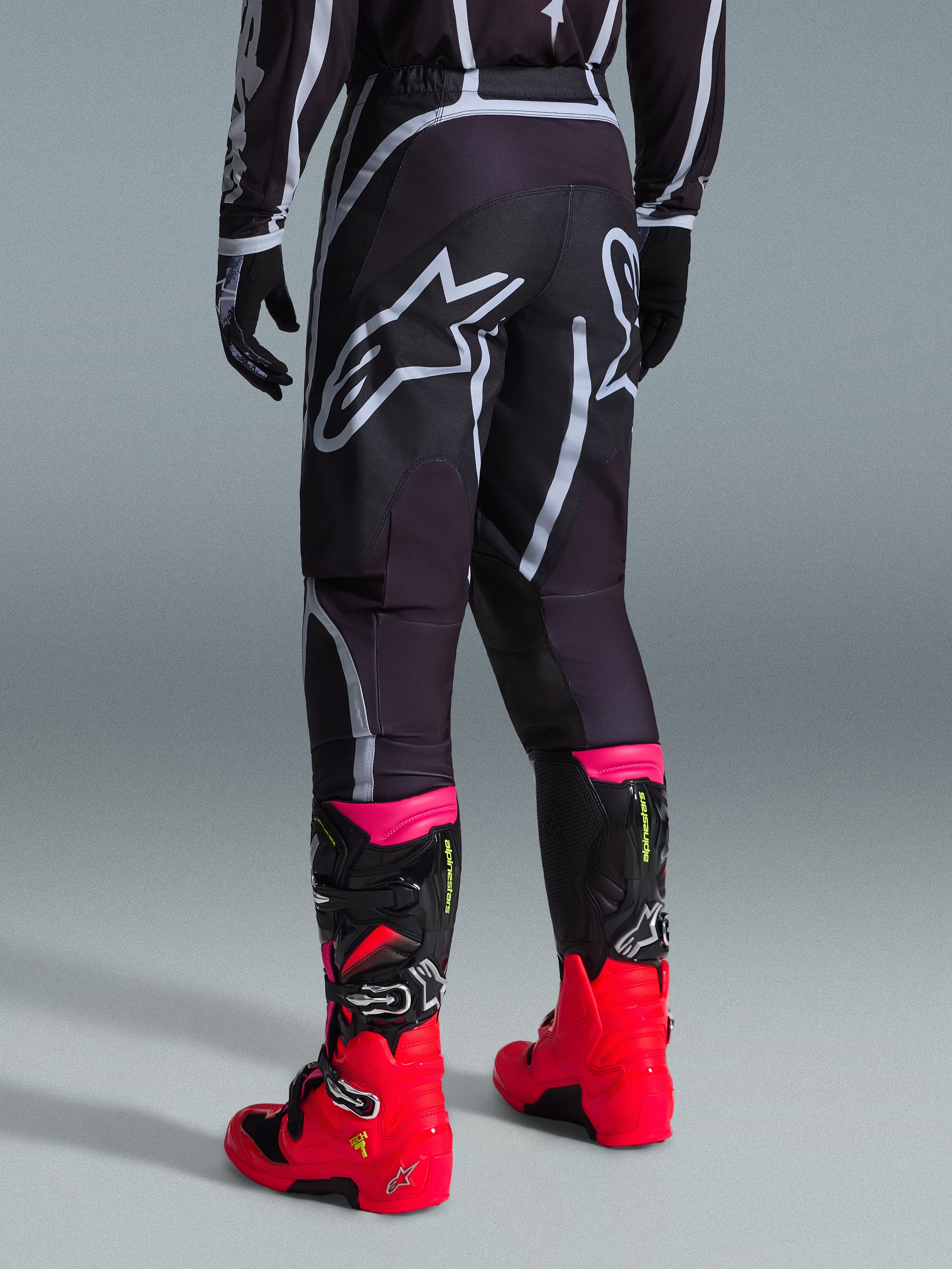 Alpinestars Pantaloni Fluid Apex 2026, pantaloni da motocross, nero e grigio, vista posteriore con grandi loghi Alpinestars bianchi sulla seduta, abbinati a stivali rosso acceso e rosa, costruzione in poliestere 600D per prestazioni off-road