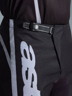 Alpinestars 2026 Fluid Apex Pants, pantaloni da motocross, nero e grigio, dettaglio ravvicinato del sistema di chiusura a cricchetto integrato per una vestibilità sicura, con grafiche del logo bianche su resistente politessuto 600D.
