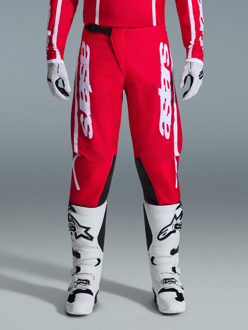 Pantaloni Alpinestars Fluid Apex 2026, pantaloni da motocross, rossi e bianchi, con grandi loghi Astars bianchi sulle cosce e pannelli interni neri sulle ginocchia, indossati con stivali e guanti coordinati bianchi e neri per un look completo da gara off-road.