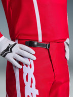 Pantaloni Alpinestars Fluid Apex 2026, pantaloni da motocross, rosso e bianco, primo piano del sistema di chiusura a cricchetto nero in vita, indossati con guanti Alpinestars bianchi e maglia rossa coordinata, equipaggiamento tecnico resistente per il fuoristrada.