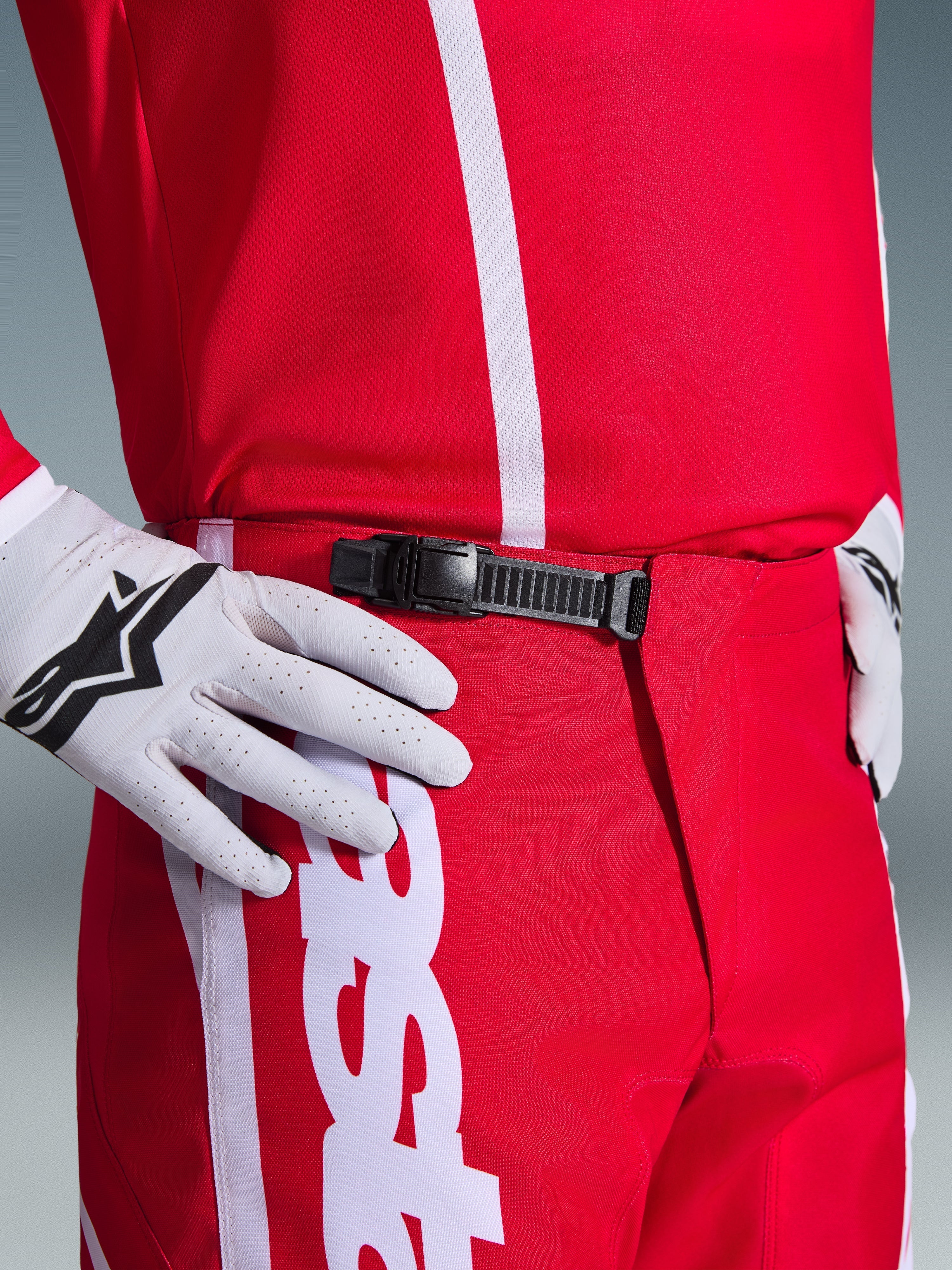 Pantaloni Alpinestars Fluid Apex 2026, pantaloni da motocross, rosso e bianco, primo piano del sistema di chiusura a cricchetto nero in vita, indossati con guanti Alpinestars bianchi e maglia rossa coordinata, equipaggiamento tecnico resistente per il fuoristrada.
