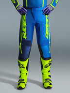 Alpinestars 2026 Fluid Apex Pants, pantaloni da motocross, blu e giallo fluo, indossati con stivali giallo fluo coordinati e guanti viola, branding giallo vivace sui lati, design ergonomico per corse fuoristrada