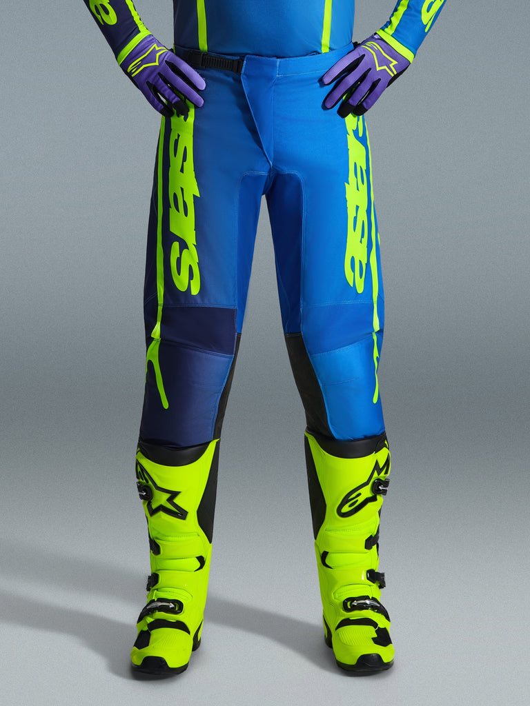 Alpinestars 2026 Fluid Apex Pants, pantaloni da motocross, blu e giallo fluo, indossati con stivali giallo fluo coordinati e guanti viola, branding giallo vivace sui lati, design ergonomico per corse fuoristrada