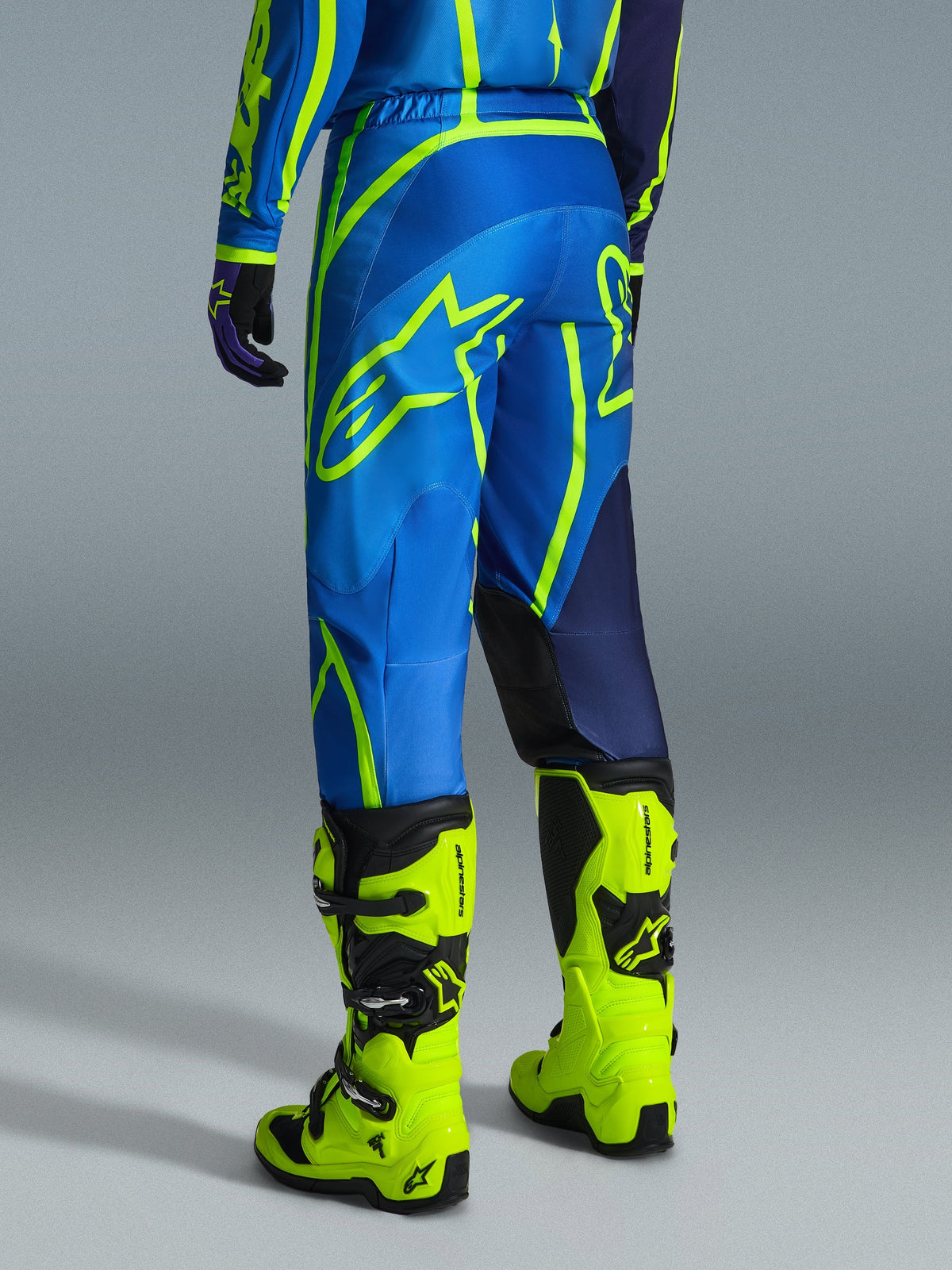 Alpinestars 2026 Fluid Apex Pants, pantaloni da motocross, blu e giallo fluo, vista posteriore del pilota con abbigliamento coordinato e stivali giallo fluorescente, caratterizzati da loghi Alpinestars e design ergonomico