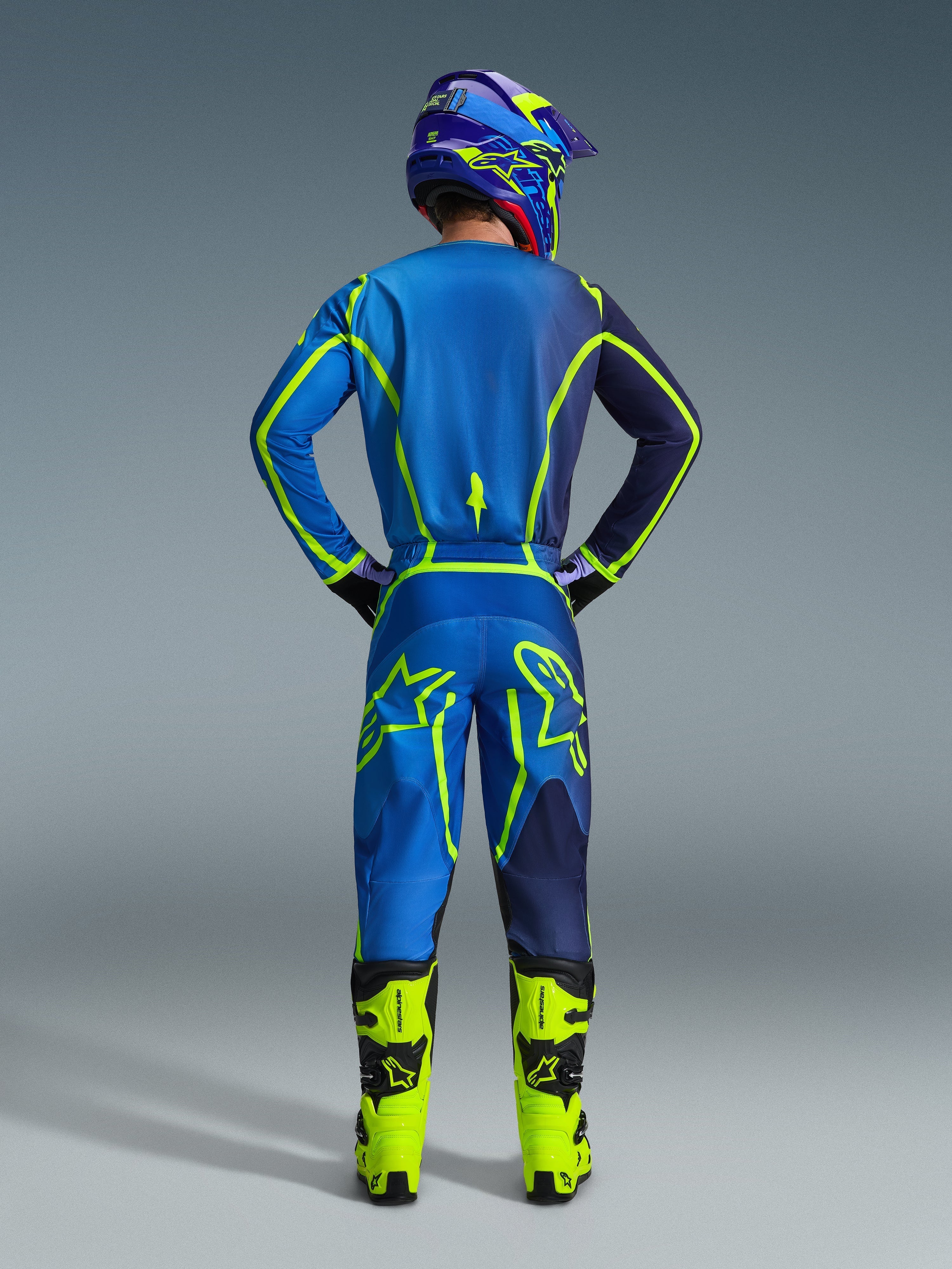 Alpinestars pantaloni Fluid Apex 2026, pantaloni da motocross, blu e giallo fluo, vista posteriore del pilota con equipaggiamento da gara completo, maglia e casco coordinati, stivali giallo brillante, vestibilità atletica coordinata per il motocross.