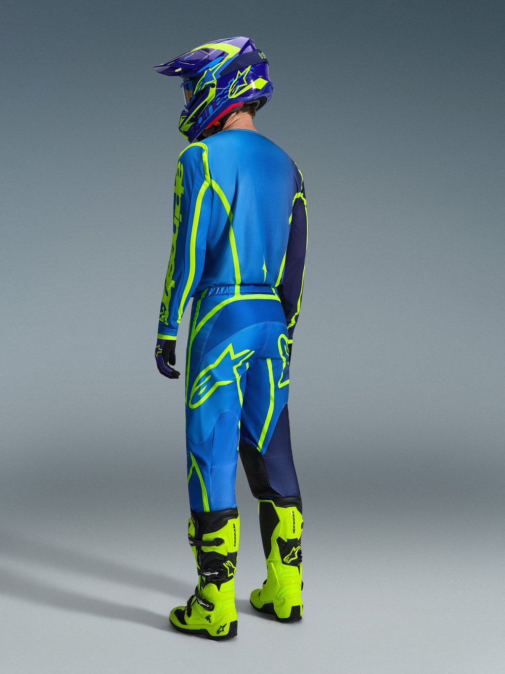 Pilota di motocross visto da dietro con pantaloni Alpinestars Fluid Apex 2026 in blu e giallo fluo, maglia e casco coordinati blu e giallo e stivali giallo neon brillante, equipaggiamento da gara coordinato per prestazioni fuoristrada