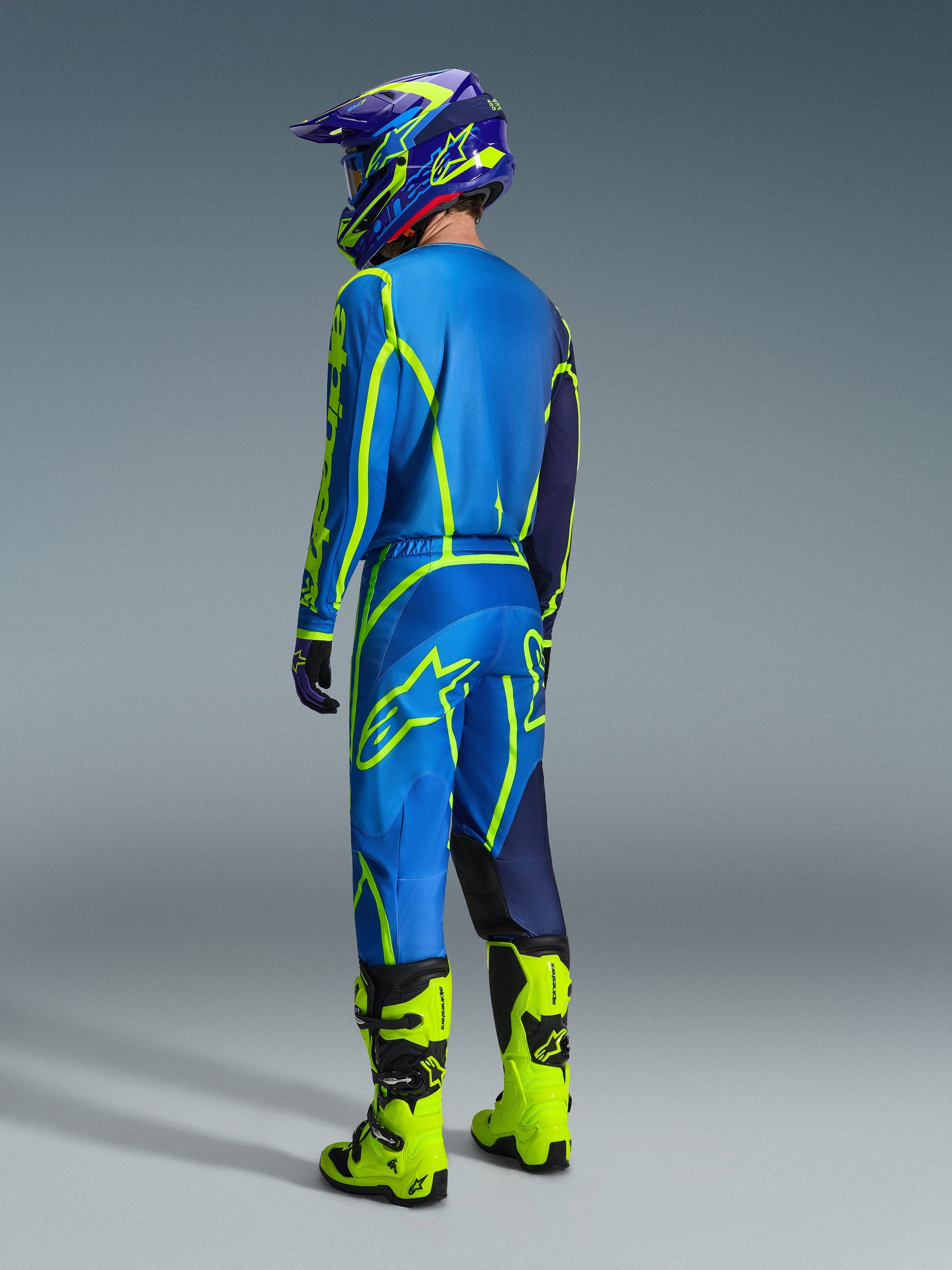 Pilota di motocross visto da dietro con pantaloni Alpinestars Fluid Apex 2026 in blu e giallo fluo, maglia e casco coordinati blu e giallo e stivali giallo neon brillante, equipaggiamento da gara coordinato per prestazioni fuoristrada