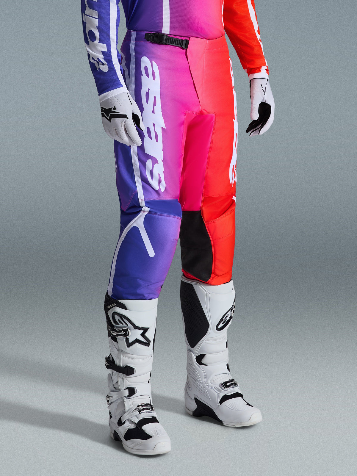 Alpinestars 2026 Fluid Apex Pants, pantaloni da motocross, sfumatura multicolore viola, rosa e rossa, indossati con stivali e guanti da motocross bianchi e neri, caratterizzati da branding verticale bianco e inserti neri sulle ginocchia, abbigliamento tecnico per fuoristrada