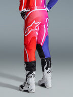 Alpinestars 2026 Fluid Apex Pants, pantaloni da motocross, multicolore rosso, rosa e viola, vista posteriore del pilota con stivali da motocross bianchi e neri, grande logo Alpinestars bianco, design sfumato vivace