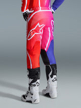 Alpinestars 2026 Fluid Apex Pants, pantaloni da motocross, multicolore rosso, rosa e viola, vista posteriore del pilota con stivali da motocross bianchi e neri, grande logo Alpinestars bianco, design sfumato vivace