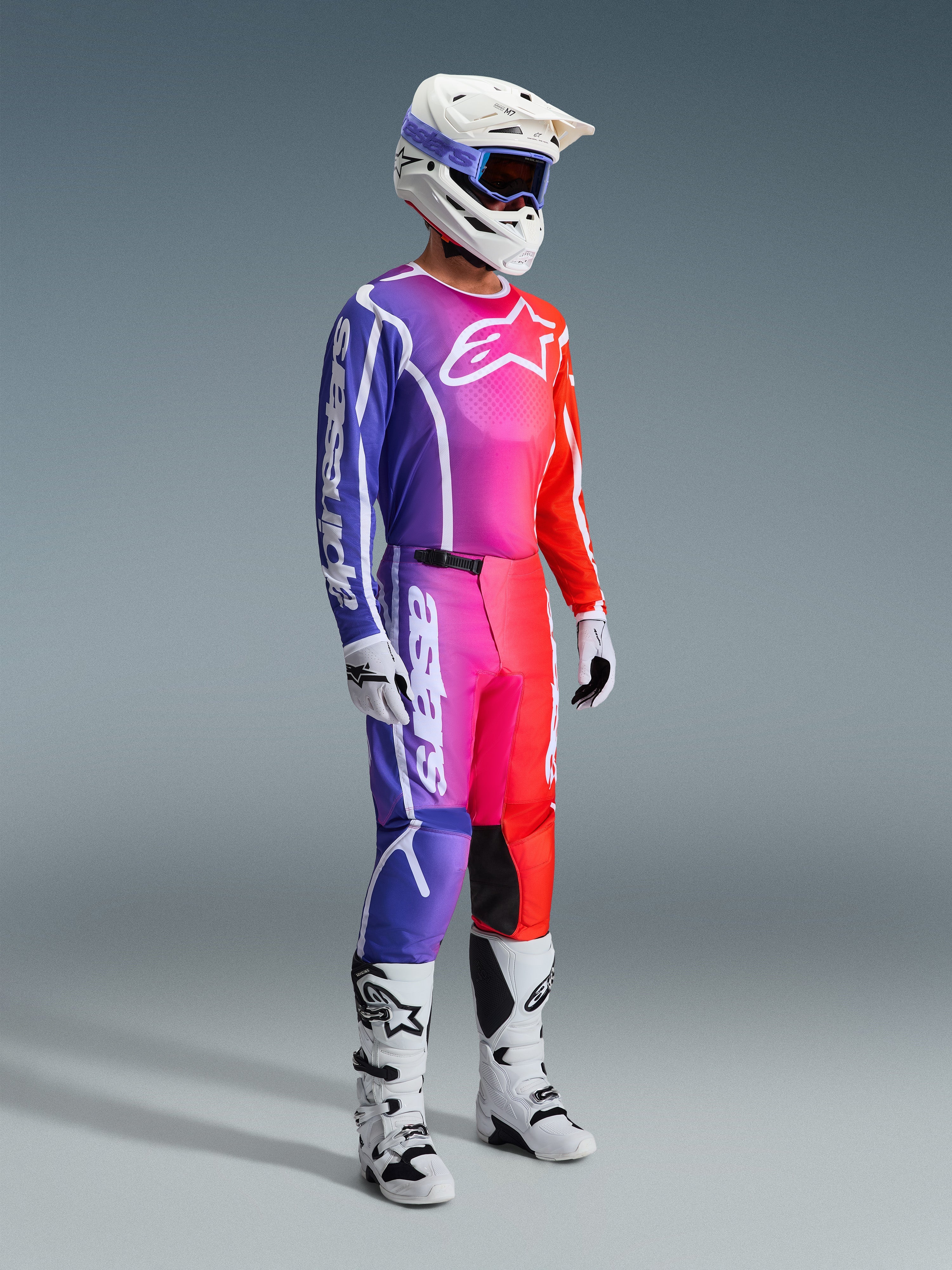 Alpinestars 2026 Fluid Apex Pants, pantaloni da motocross, sfumatura multicolore viola, rosa e arancione, indossati con maglia abbinata, casco bianco e stivali bianchi, con logo bianco e design ergonomico per corse off-road