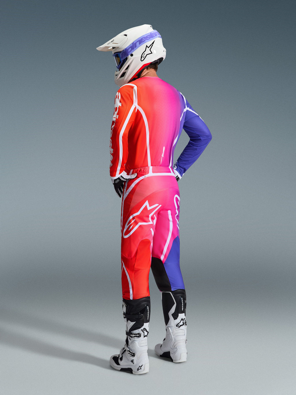 Pilota di motocross con abbigliamento Alpinestars 2026 Fluid Apex, maglia e pantaloni multicolore con una sfumatura rossa, rosa e viola e loghi bianchi, casco bianco e stivali bianchi, outfit coordinato per gare fuoristrada