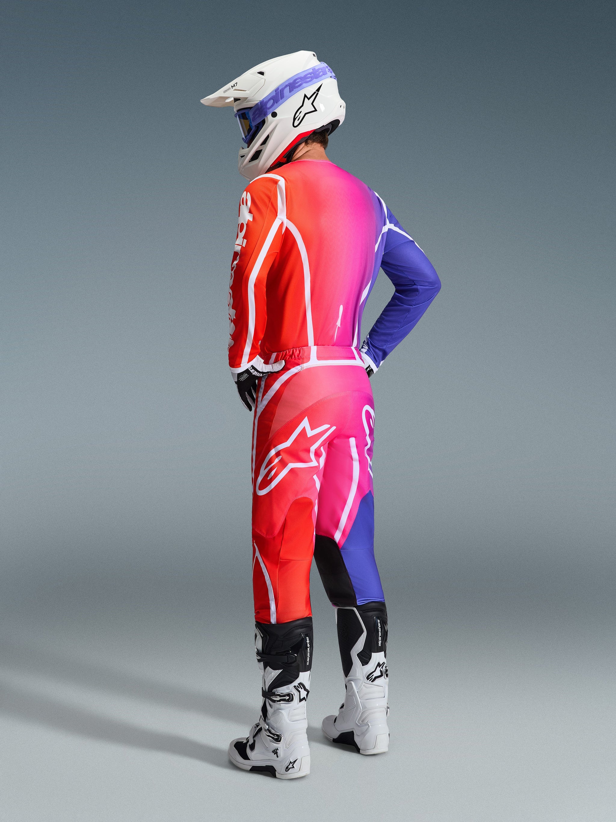 Pilota di motocross con abbigliamento Alpinestars 2026 Fluid Apex, maglia e pantaloni multicolore con una sfumatura rossa, rosa e viola e loghi bianchi, casco bianco e stivali bianchi, outfit coordinato per gare fuoristrada