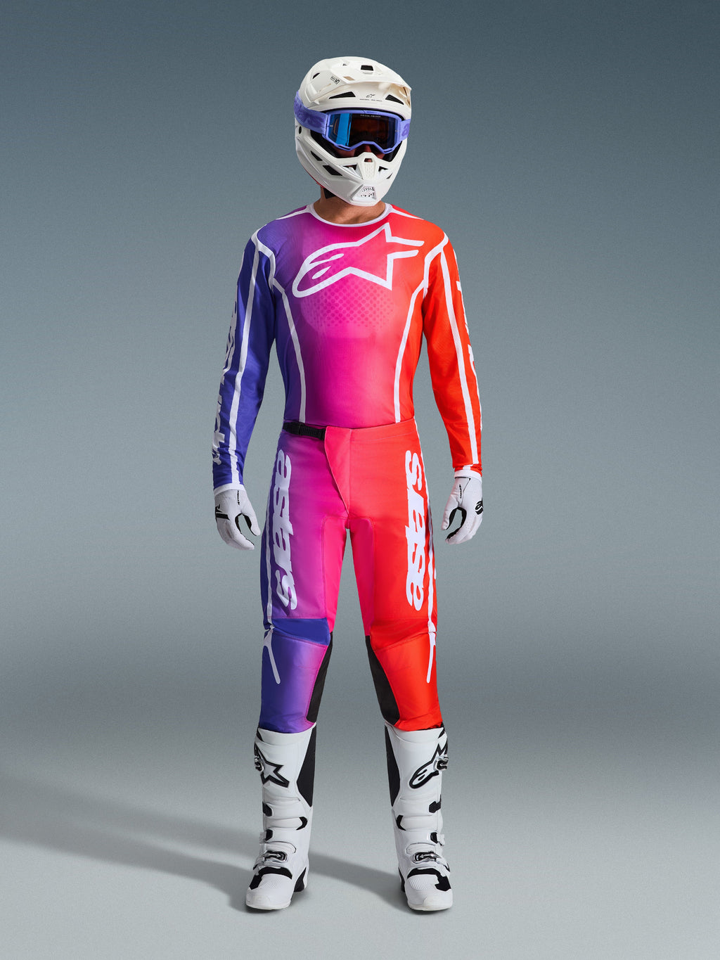 Pilota di motocross con pantaloni Fluid Apex 2026 e maglia coordinata, sfumatura viola e arancione, casco bianco con maschera, stivali e guanti bianchi, abbigliamento da gara Alpinestars coordinato per prestazioni off-road.