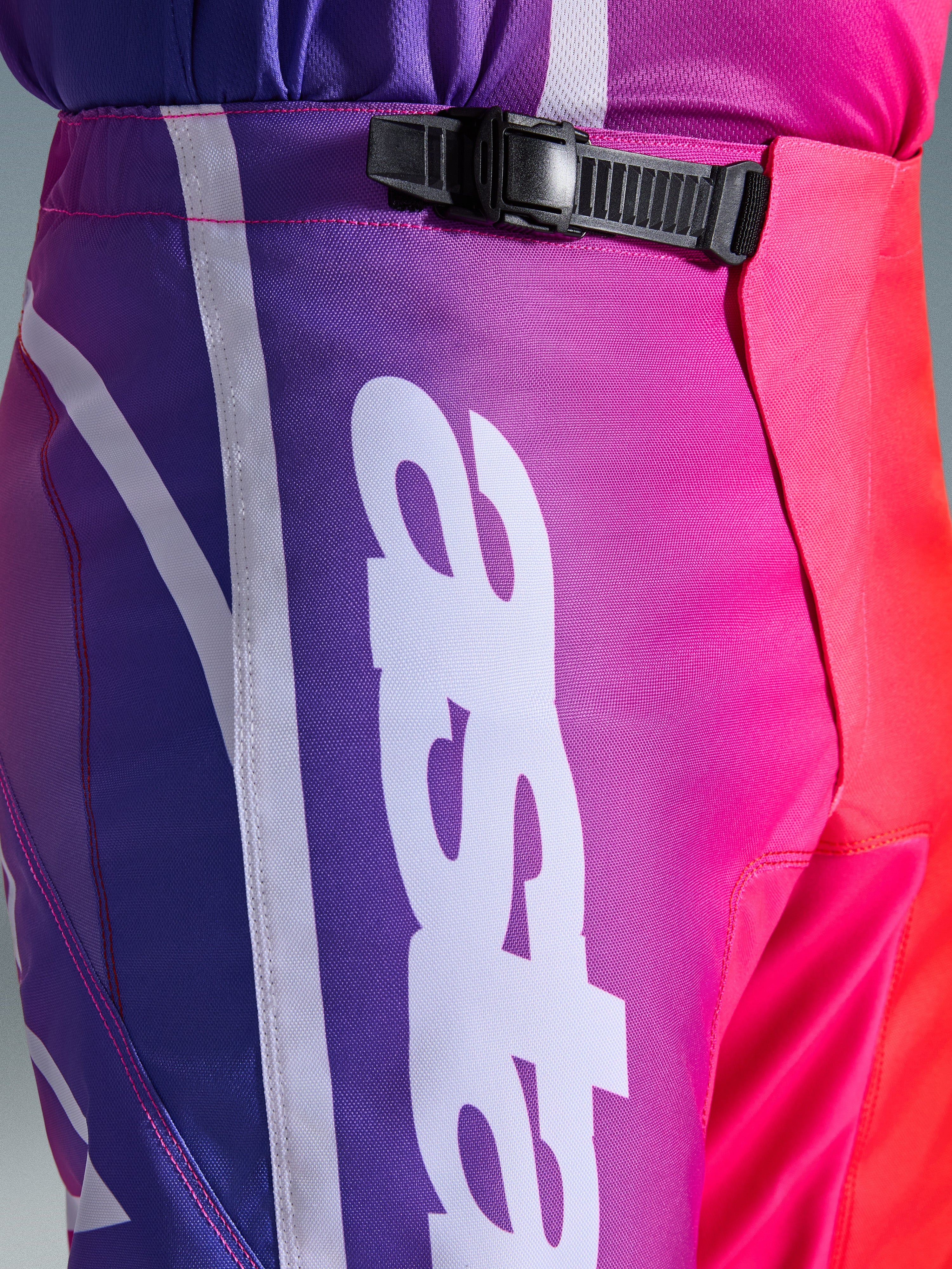 Alpinestars 2026 Fluid Apex Pants, pantaloni da motocross, multicolore viola, rosa e rosso, primo piano della zona vita e fianchi con sistema di chiusura a cricchetto nero e grande logo Alpinestars verticale bianco, design sfumato vivace con trama in tessuto poliestere resistente
