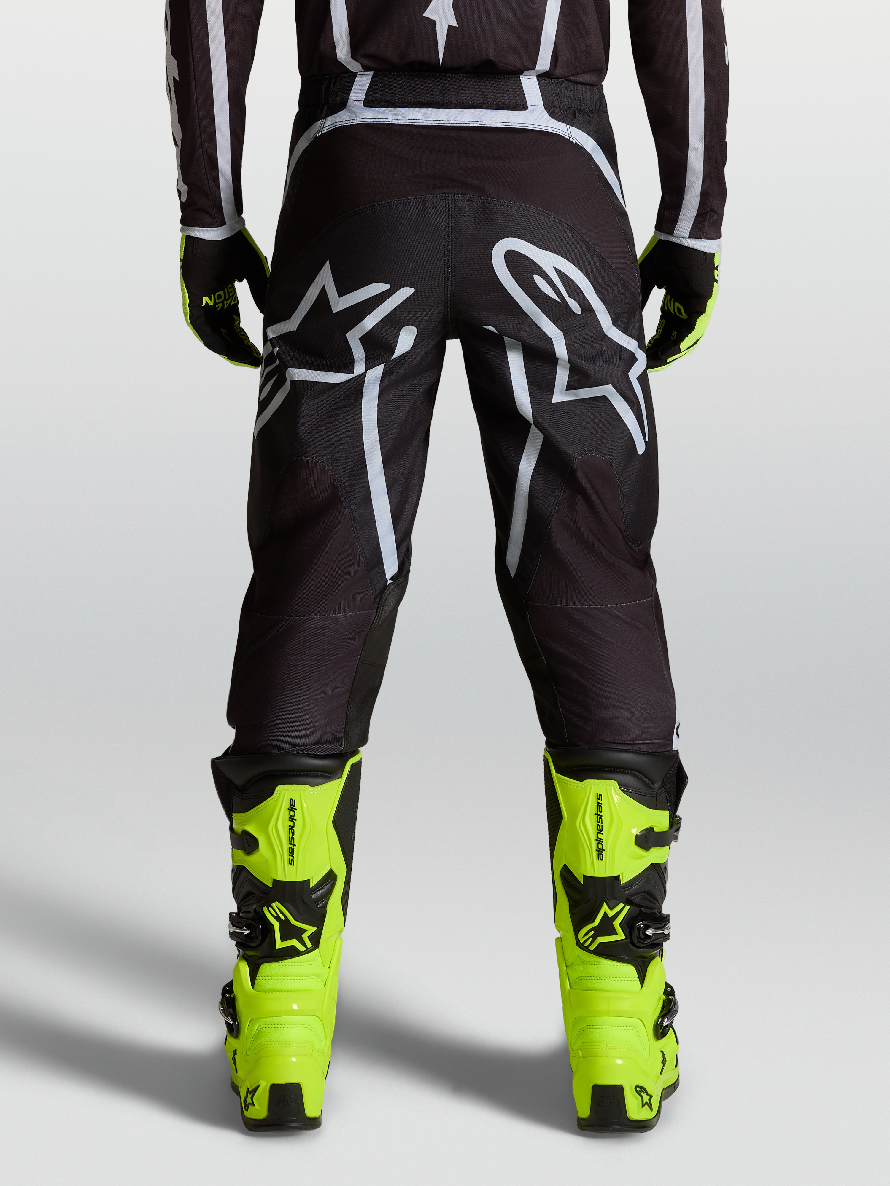 Alpinestars Pantaloni Fluid Apex V2, pantaloni motocross da uomo, nero e argento, vista posteriore con grandi loghi Astars e dettagli in maglia mesh, indossati con stivali giallo fluo per performance fuoristrada