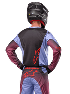 Pilota di motocross visto di spalle con abbigliamento Alpinestars Racer Hoen 2024, maglia Lavan Purple e nera con logo rosso, pantaloni coordinati con dettagli rossi, casco nero, completo coordinato per corse fuoristrada