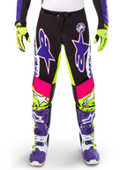 Alpinestars Limited Edition Supertech Dirt Studios Pants, pantaloni da motocross, bianco, viola e giallo fluo, con pannelli neri e accenti giallo neon e rosa, grandi loghi a stella viola, mostrati con guanti giallo neon e stivali da corsa viola coordinati, abbigliamento da corsa fuoristrada resistente