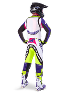 Pilota di motocross che indossa pantaloni Alpinestars Limited Edition Supertech Dirt Studios® in Bianco, Viola e Giallo Fluo, visto da dietro con maglia e stivali coordinati e casco in carbonio nero, abbigliamento professionale da gara off-road con grafiche audaci e accenti neon.