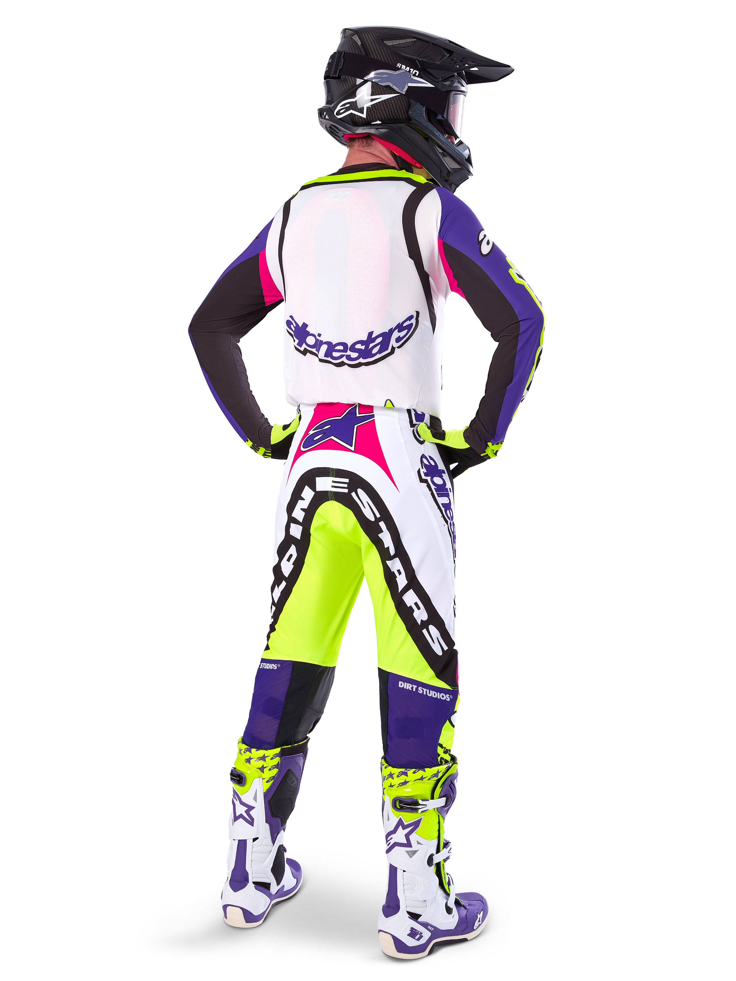 Pilota di motocross che indossa pantaloni Alpinestars Limited Edition Supertech Dirt Studios® in Bianco, Viola e Giallo Fluo, visto da dietro con maglia e stivali coordinati e casco in carbonio nero, abbigliamento professionale da gara off-road con grafiche audaci e accenti neon.